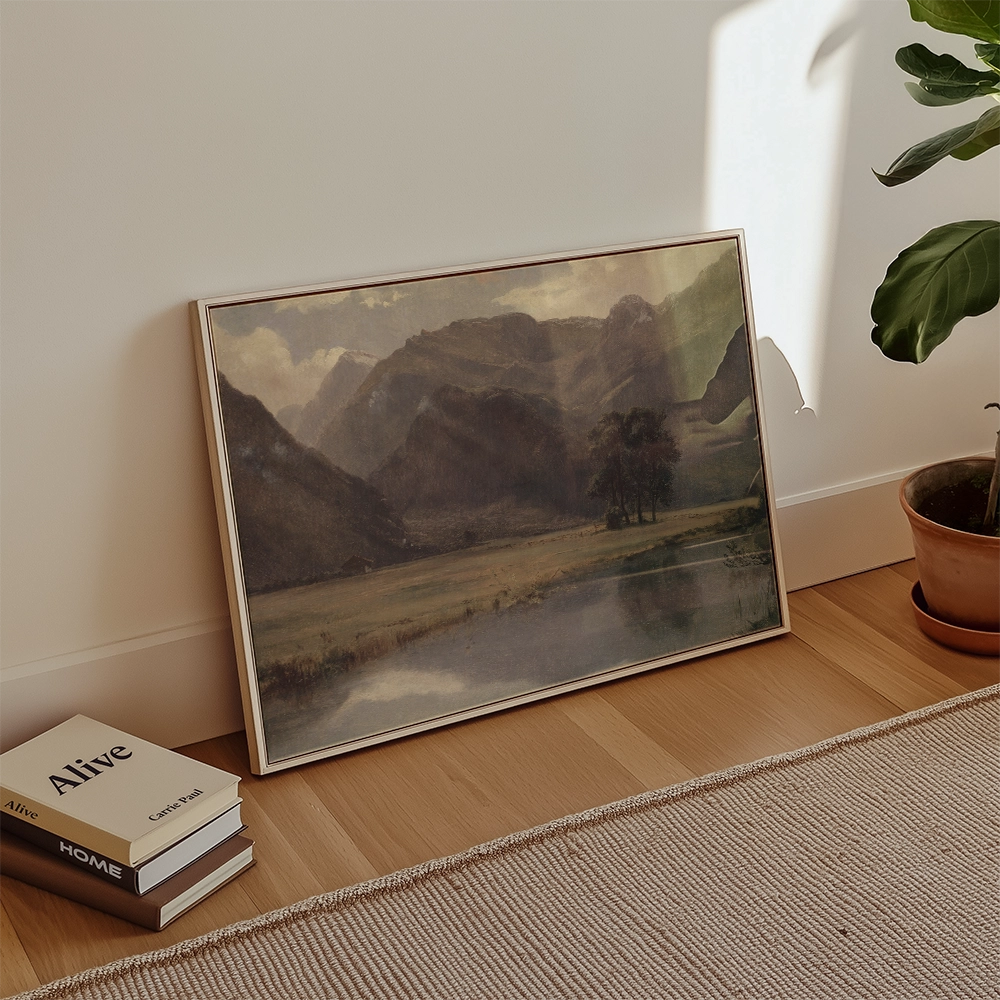 Framed landscape painting featuring mountains and a serene lake, positioned on a wooden floor beside stacked books and a potted plant.