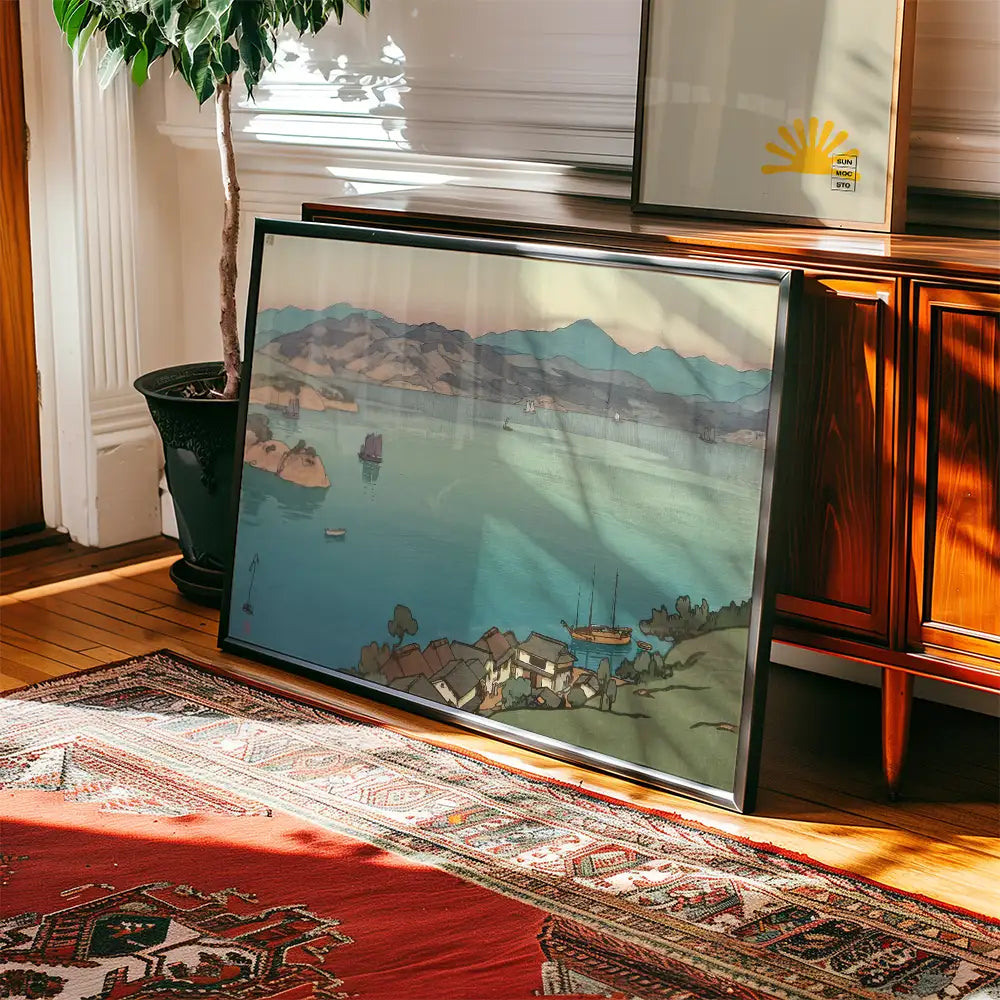 Framed landscape painting featuring a serene lake with mountains in the background, placed on a wooden floor next to a plant and an ornate rug.
