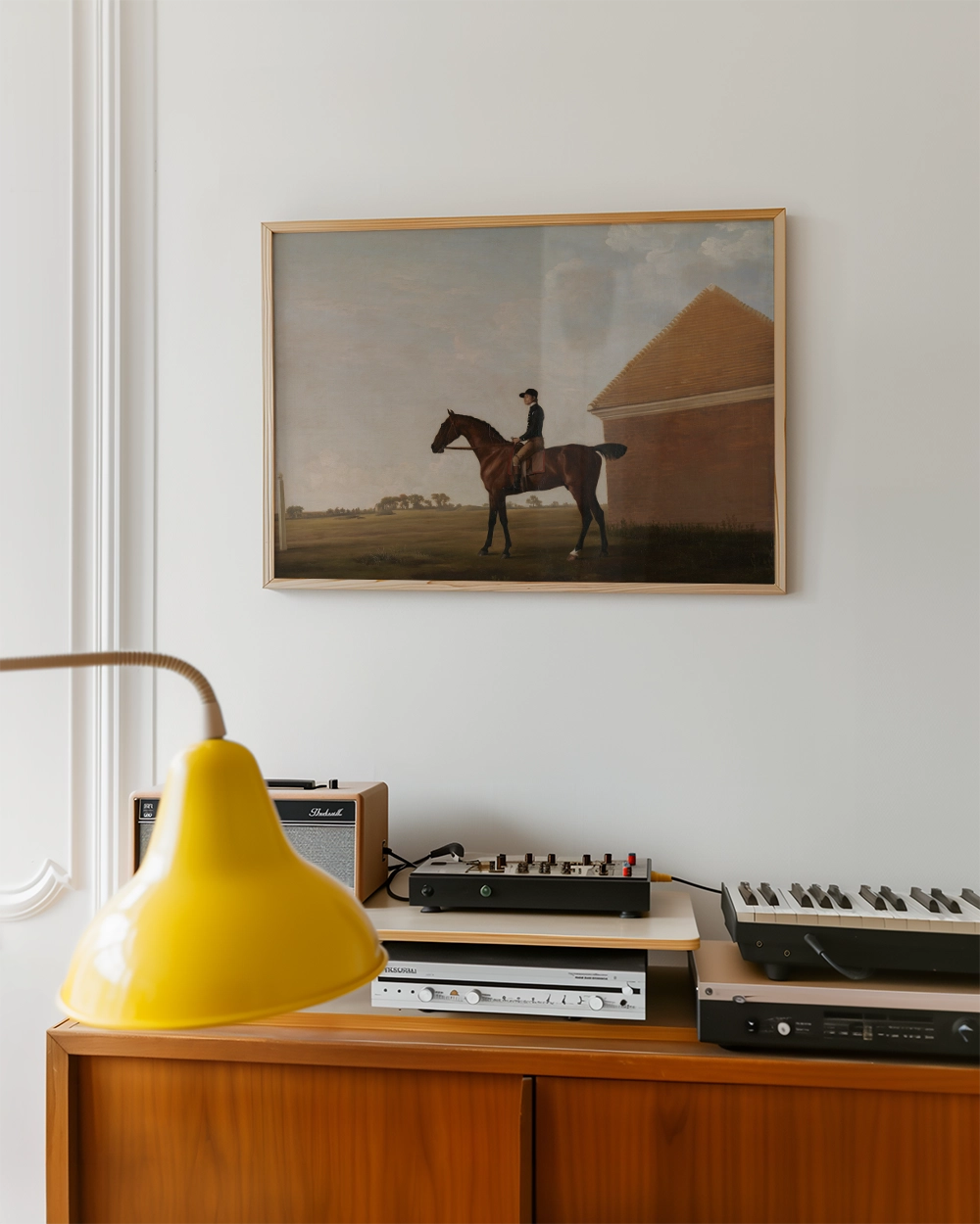 Painting of a horse and rider hanging on the wall above a wooden console table with a yellow lamp and various audio equipment.