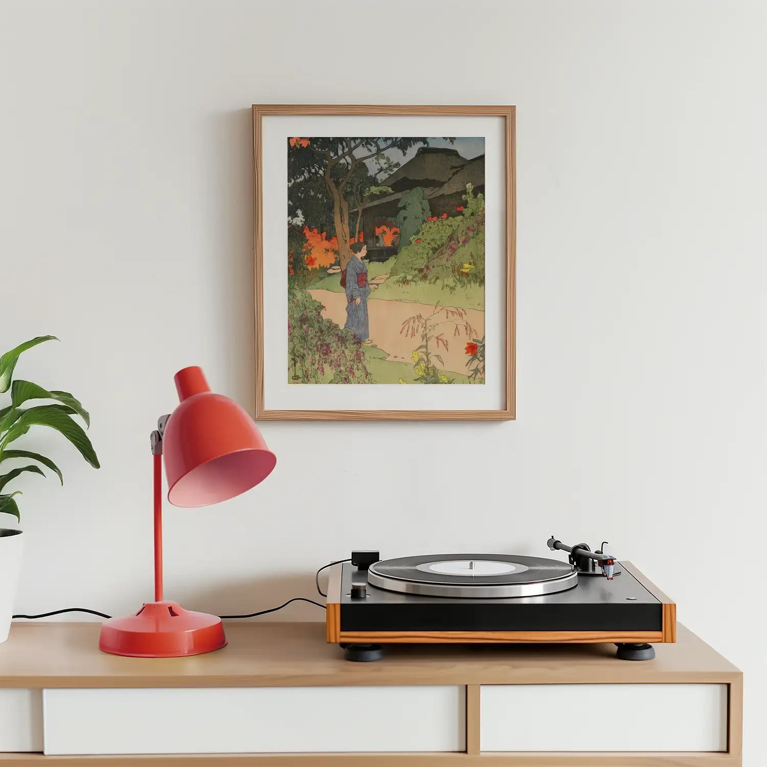 Framed painting of a serene landscape featuring a person walking along a path surrounded by vibrant flowers, displayed on a white wall above a record player and a red desk lamp.