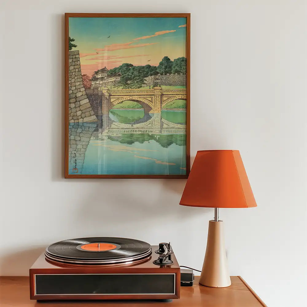 Vintage landscape painting of a bridge reflected in water, framed on a wall next to a turntable and an orange lampshade.