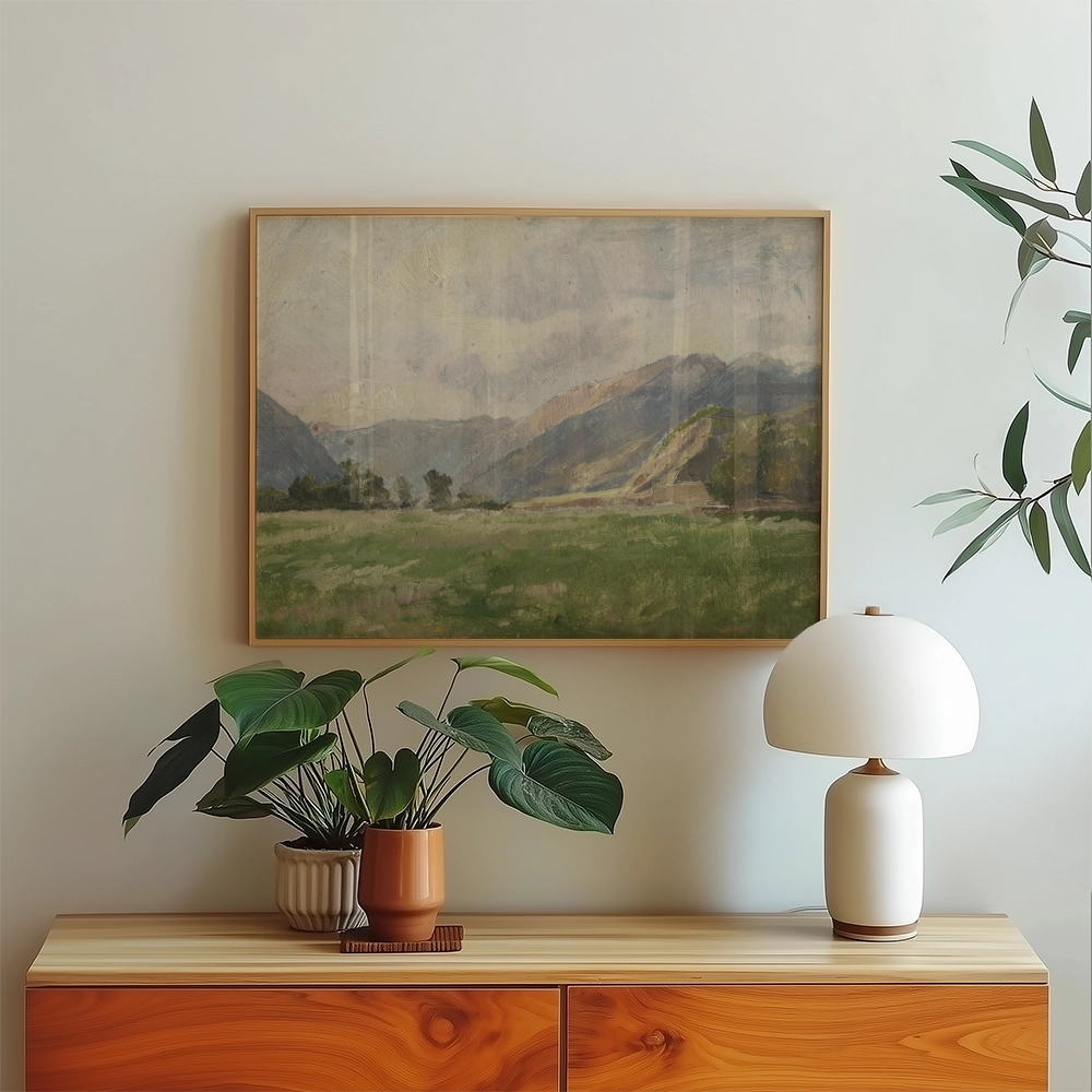 Framed painting of a serene landscape featuring mountains and grassy fields, displayed above a wooden console table with decorative plants and a modern lamp.