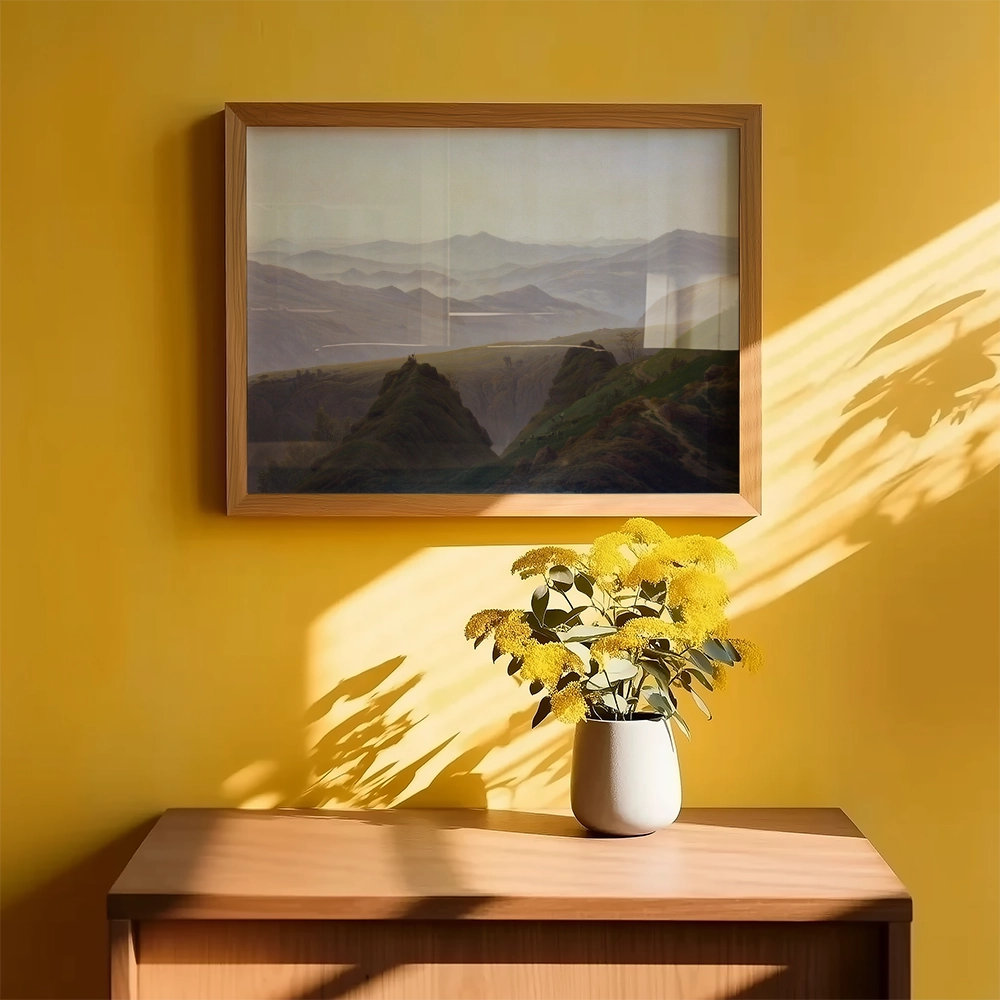 Framed landscape painting on a yellow wall above a wooden table with a vase of yellow flowers.
