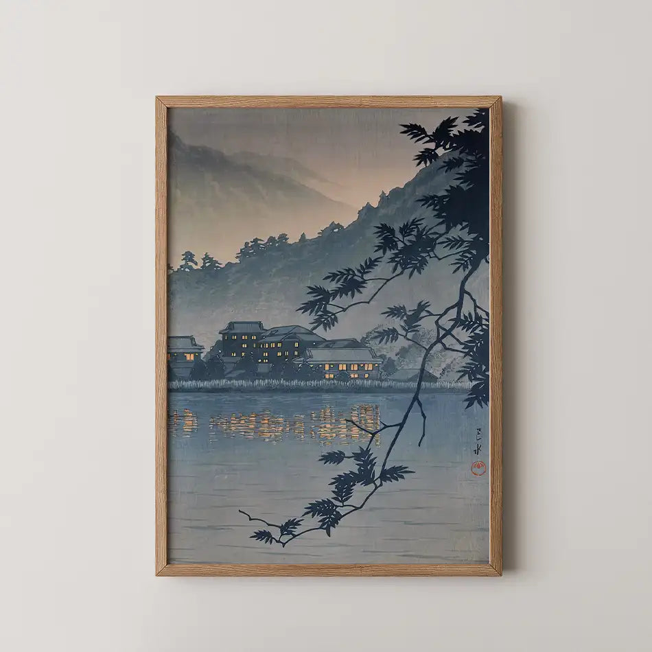 Framed painting of a serene landscape featuring a calm lake reflecting illuminated buildings and mountains under a dusk sky, with silhouetted branches in the foreground.