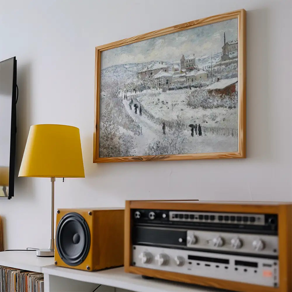 Painting of a snowy landscape with figures walking along a path, framed in wood, displayed on a wall with a yellow lamp and vintage audio equipment nearby.
