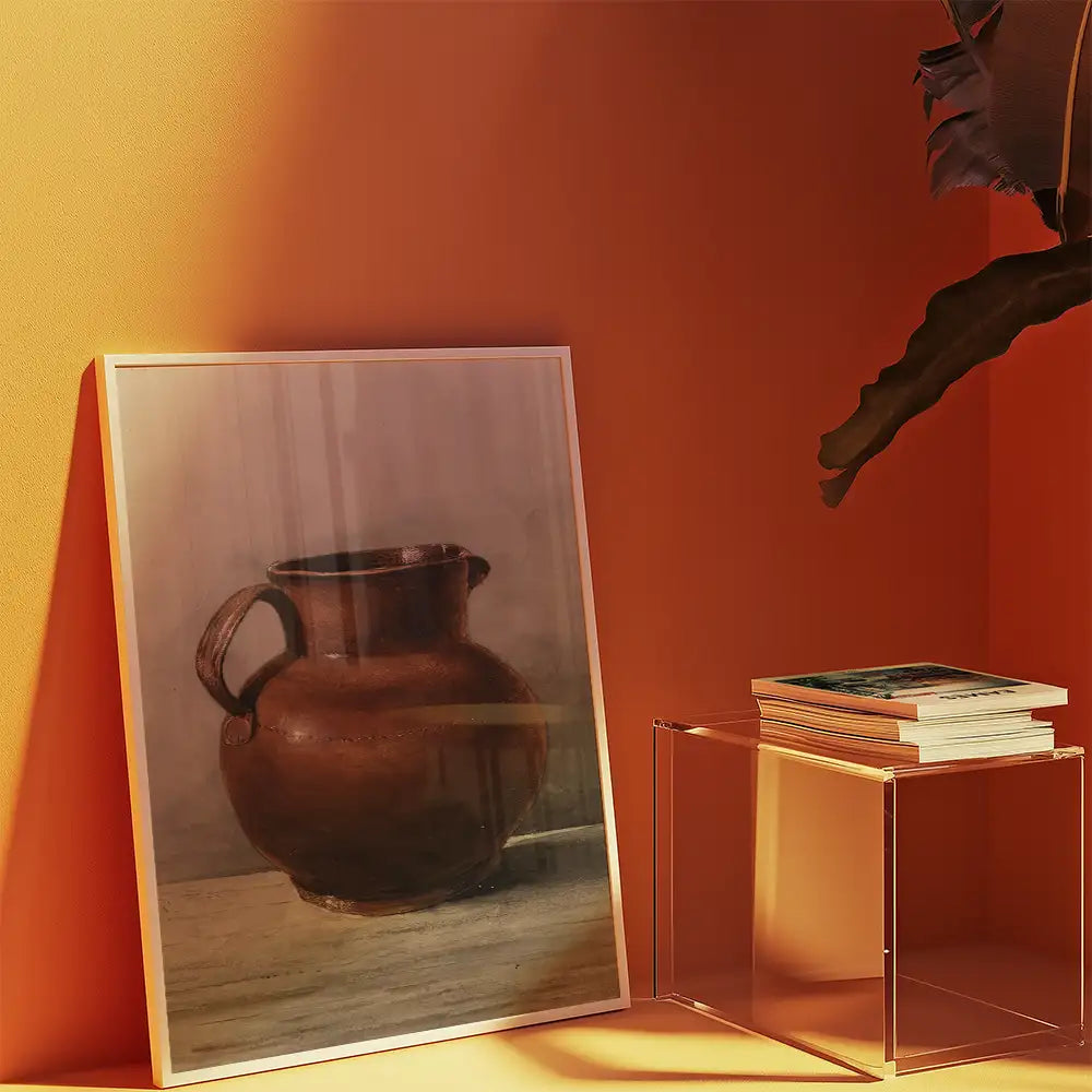 Framed painting of a brown pottery jug displayed against a textured orange wall, with a clear acrylic side table holding stacked books beside it.