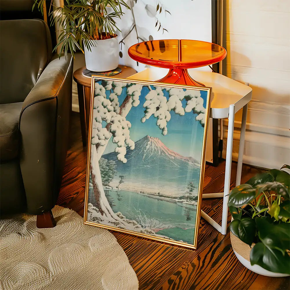 Framed painting of a snow-covered landscape featuring a mountain and a lake, placed beside a chair and table in a cozy interior setting.