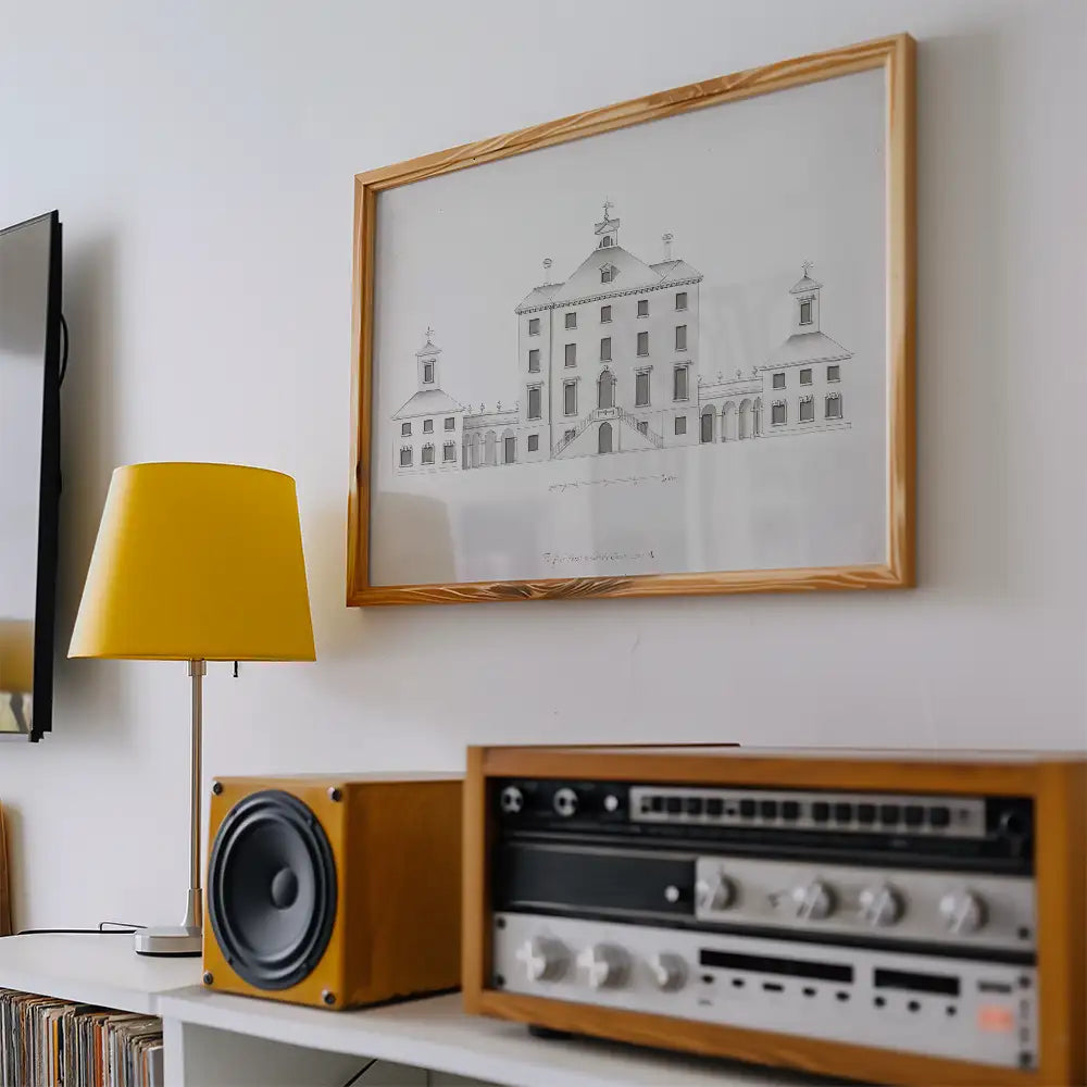 Framed architectural drawing of a building on a white wall, accompanied by a yellow lamp and wooden speakers.