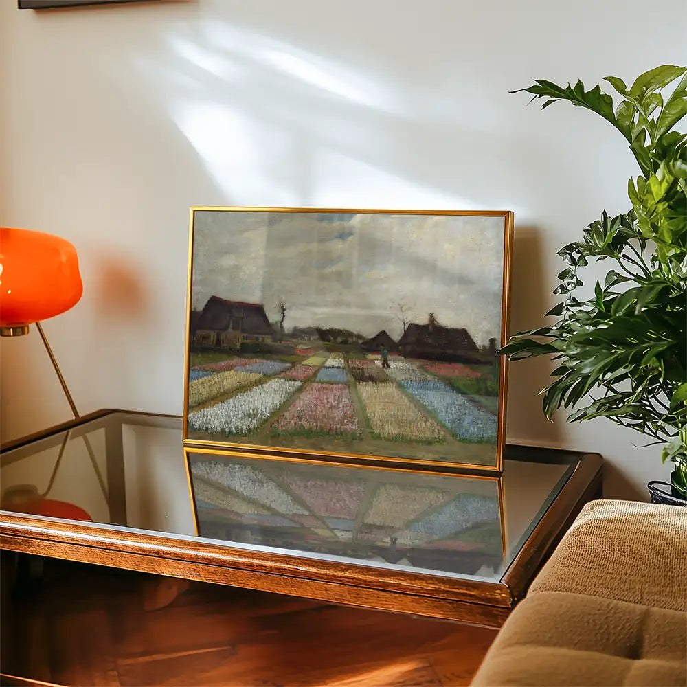 A framed painting of a landscape with colorful rows of flowers, set on a glass table next to a vintage orange lamp and a green plant.