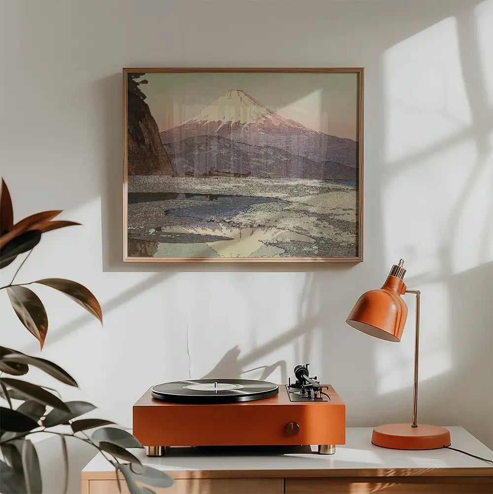 Framed painting of a mountain landscape above a vintage record player and a modern orange desk lamp in a bright room.