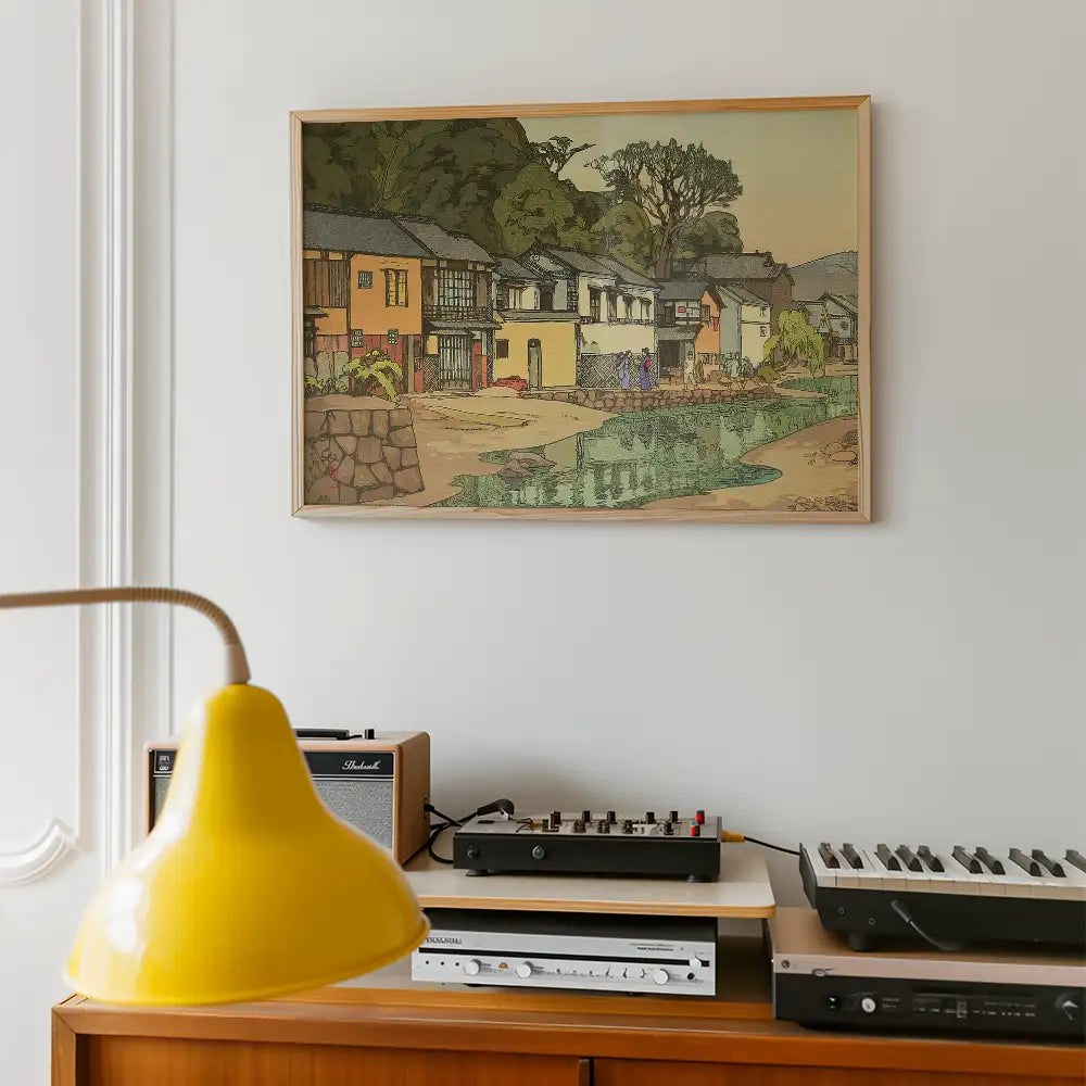Framed painting of a quaint village scene featuring colorful houses alongside a river, displayed on a wall above a wooden console with a yellow lamp and audio equipment.