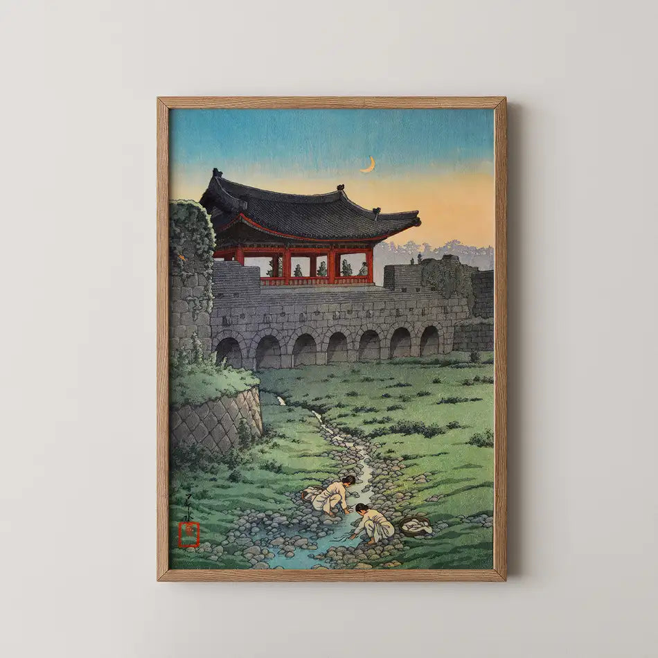 Framed painting depicting a serene landscape with a traditional building, a flowing stream, and two figures washing clothes in a grassy field at dusk.