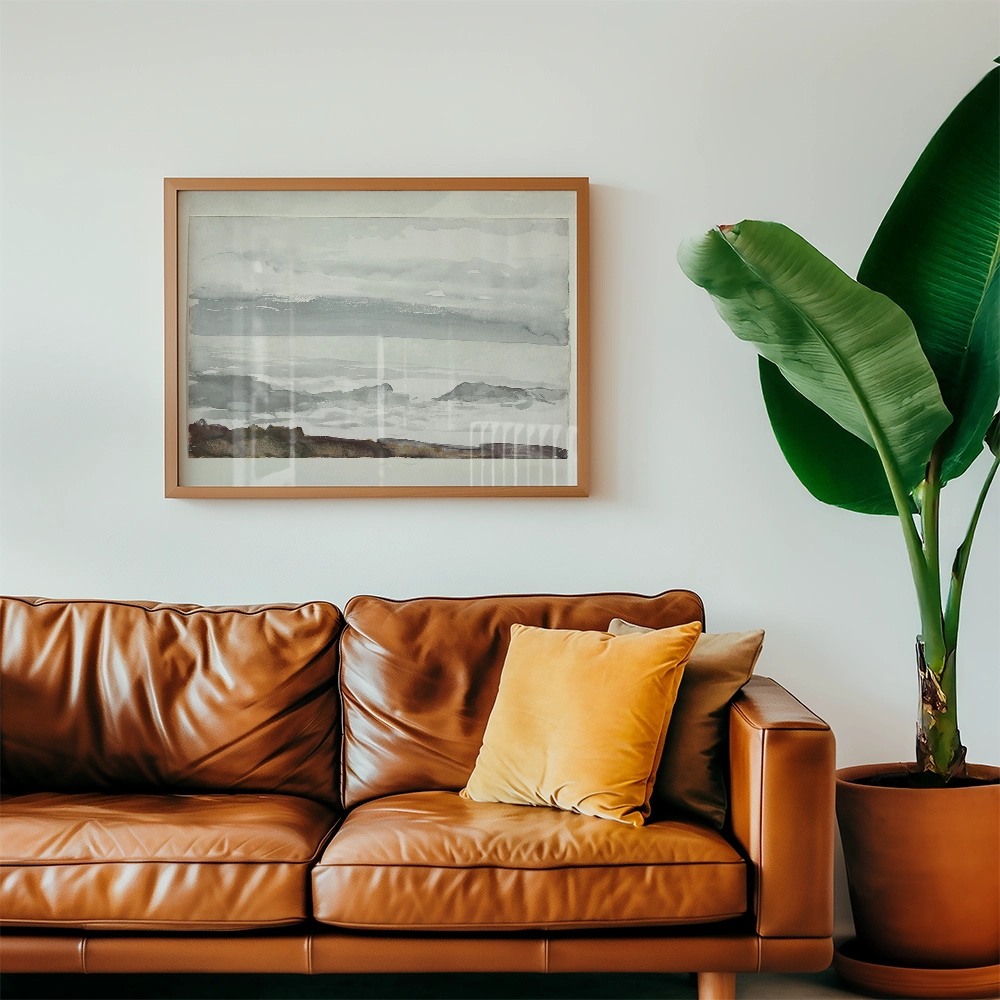 Framed watercolor painting of a serene landscape hanging on a wall above a brown leather sofa with cushions and a large green plant nearby.