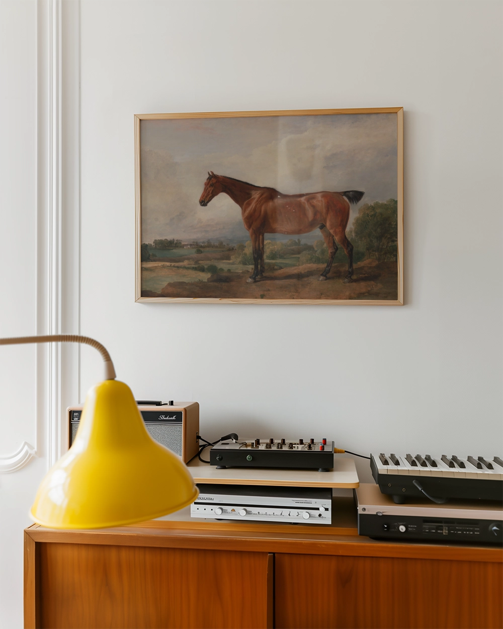 Painting of a brown horse framed on a wall above a wooden side table with a yellow lamp and audio equipment.