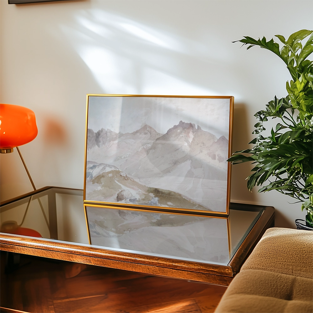 Framed mountain landscape painting displayed on a glass-top table with an orange lamp and green plant in a cozy interior setting.