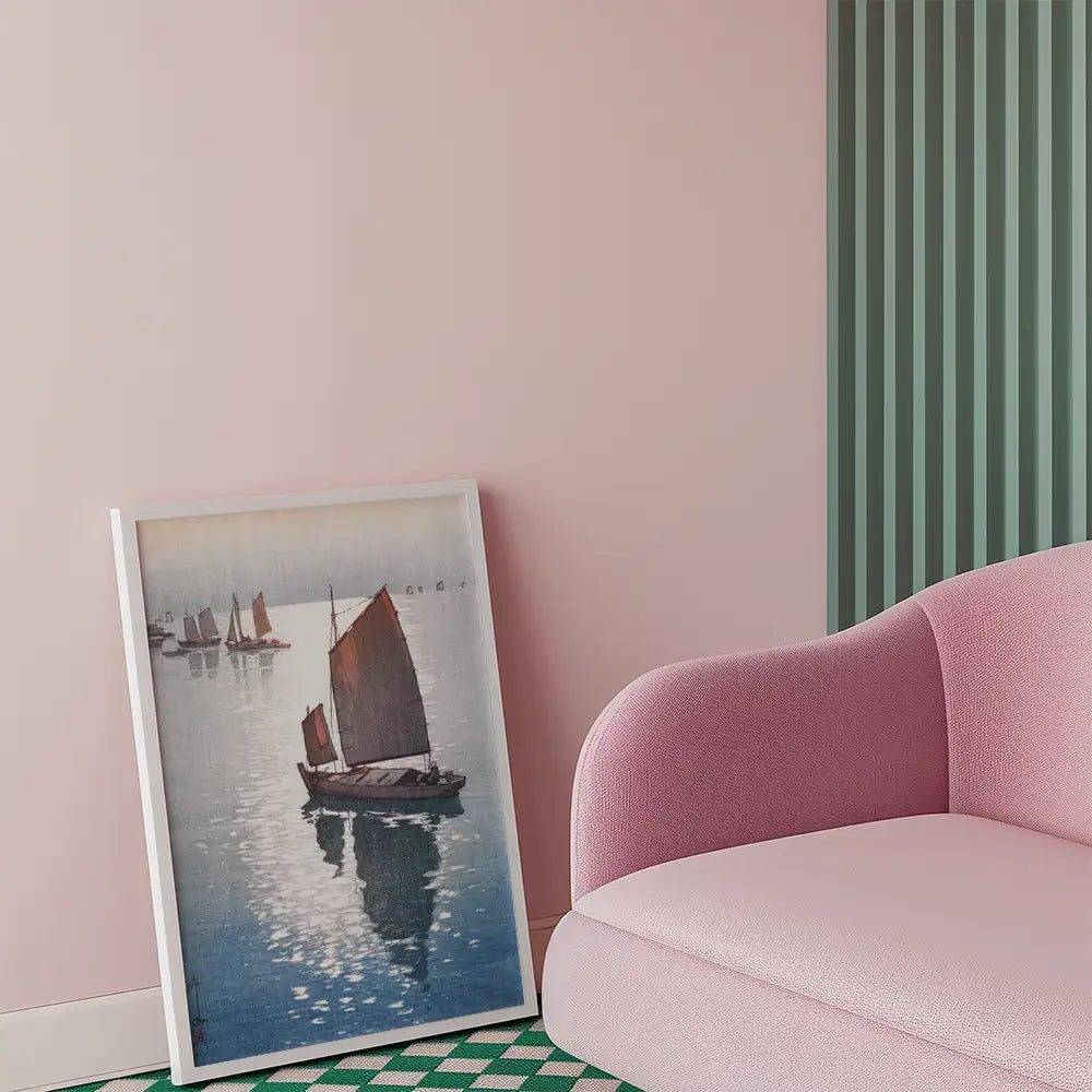 Framed painting of sailing boats on calm water leaning against a pink wall next to a modern pink sofa.