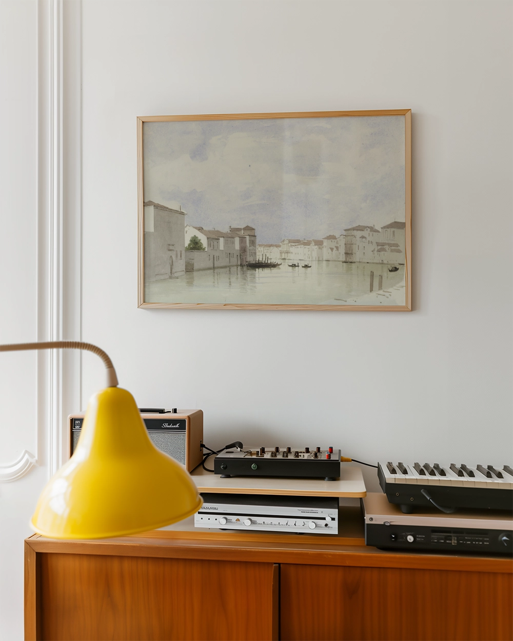 Watercolor painting of a serene canal scene framed and hung on a wall, above a wooden console with a yellow lamp and audio equipment.