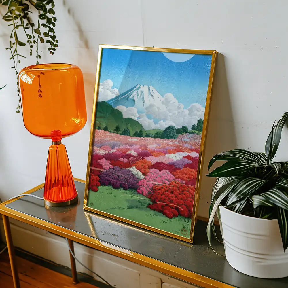Framed painting of a vibrant landscape with colorful flowers and a mountain in the background, displayed on a table beside an orange lamp and a potted plant.