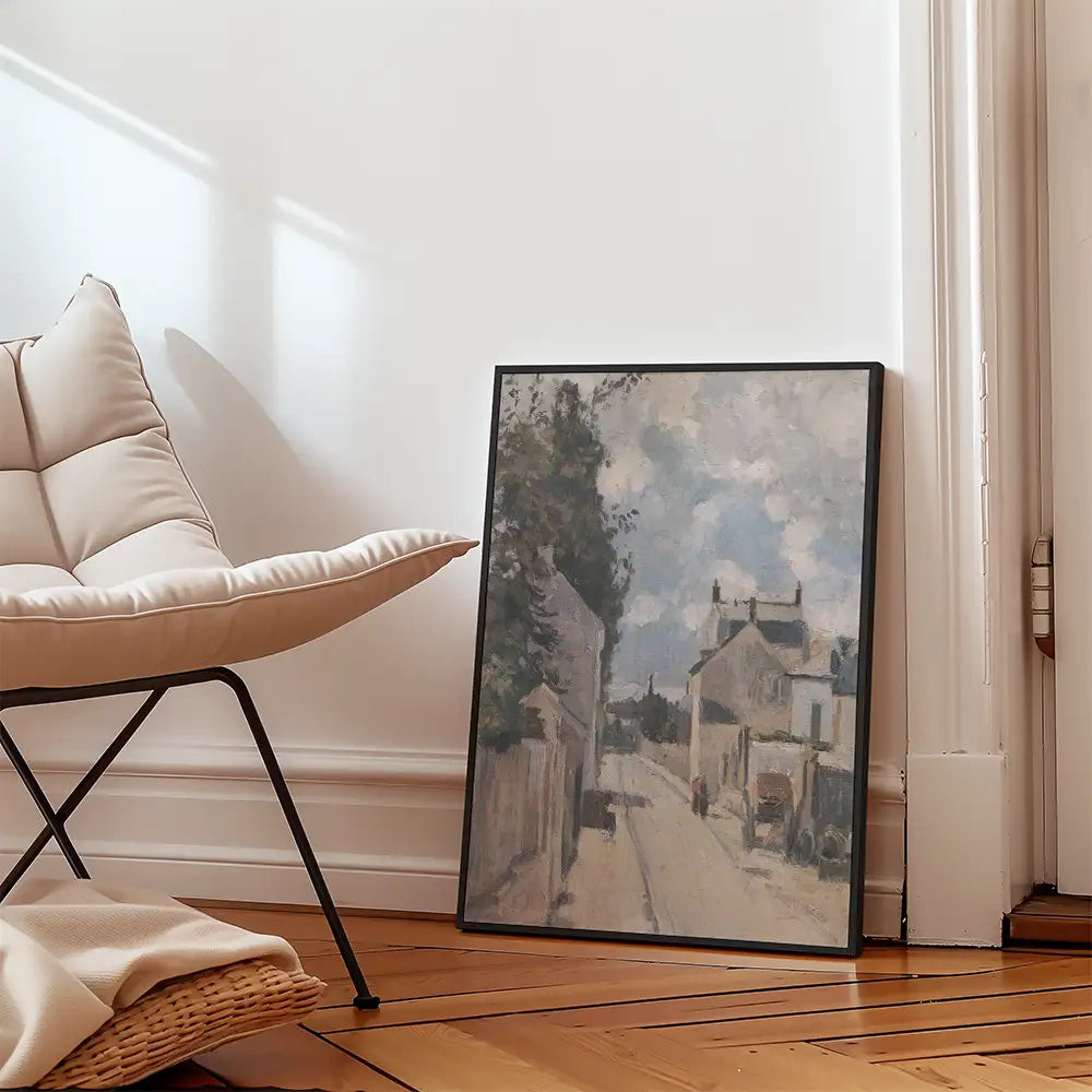 A framed painting of a scenic street view leaning against a wall next to a modern chair with soft cushions.