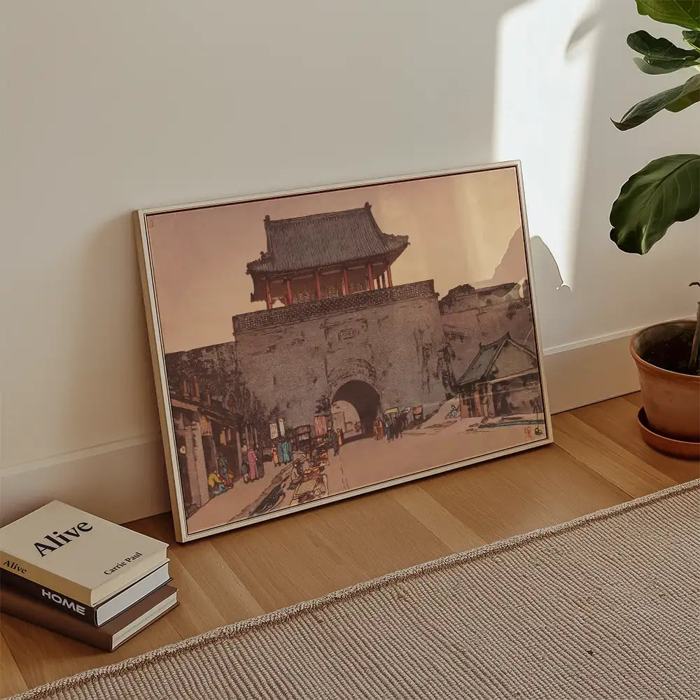 Framed painting of an ancient gate with people walking in front, displayed on a wooden floor beside stacked books and a potted plant.