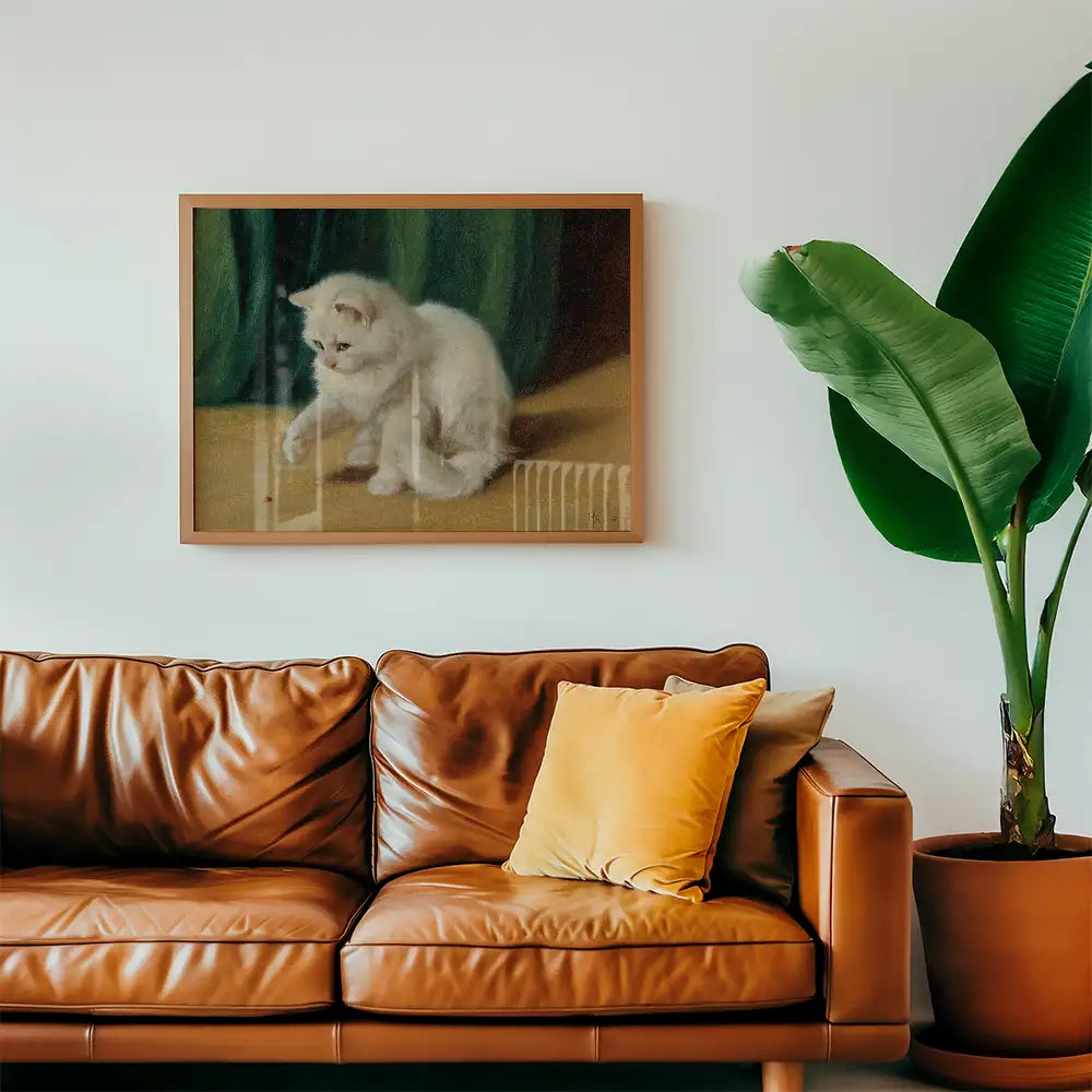 Painting of a white kitten sitting, framed and hanging on a wall above a brown leather couch with yellow pillows and a green plant nearby.