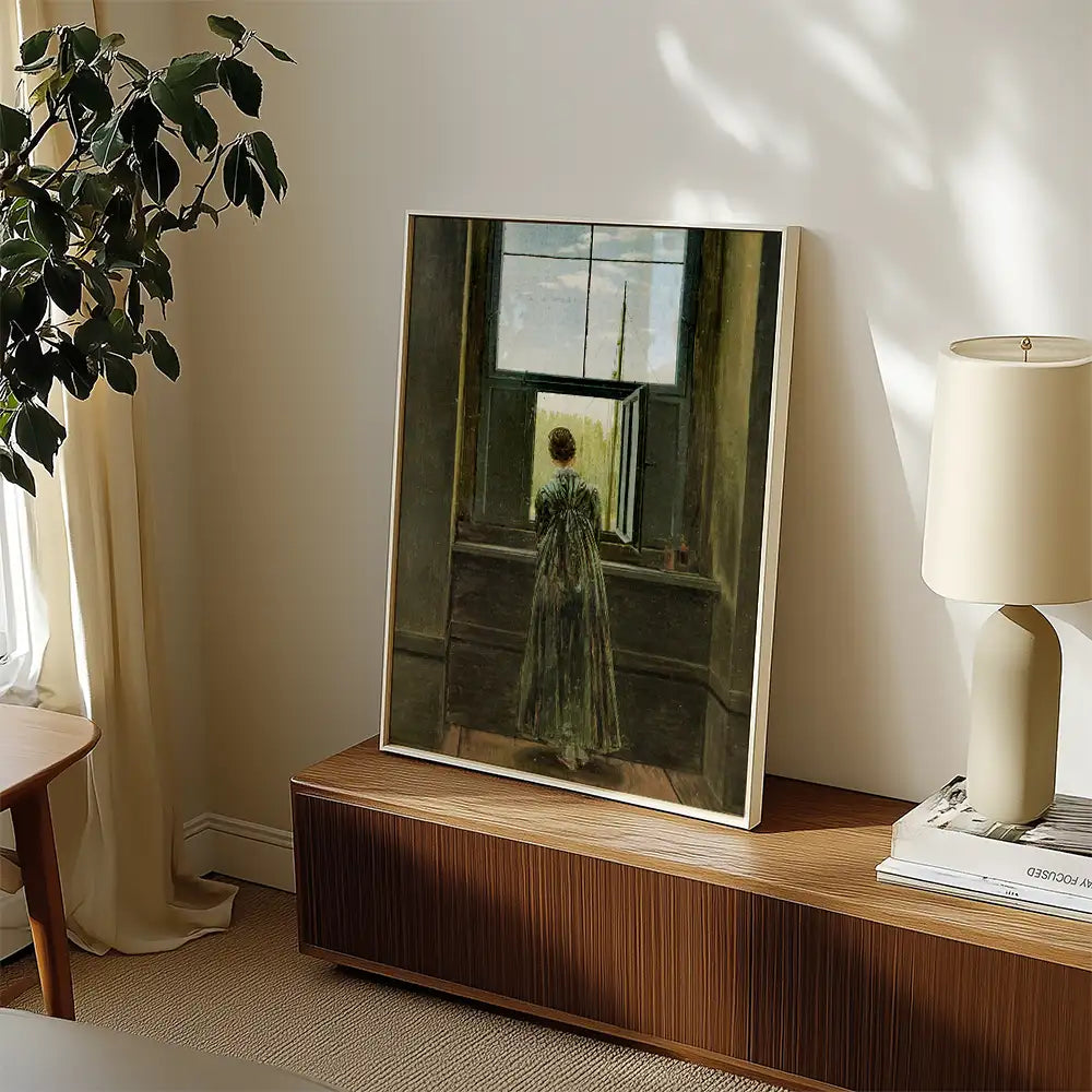 A woman in a long green dress standing by an open window, gazing at a painting in a well-lit room with modern decor.