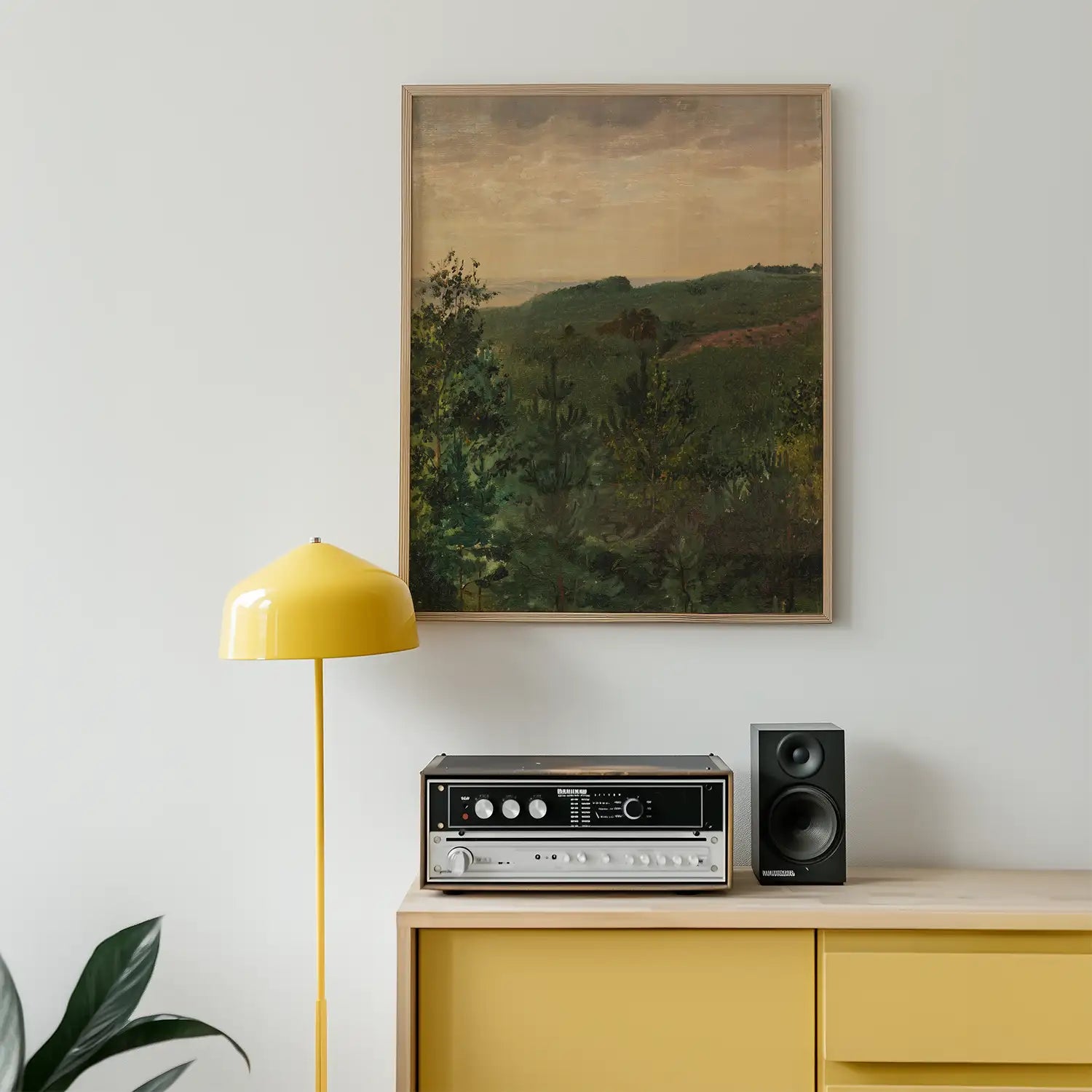 A framed landscape painting of green hills and trees hanging on a white wall, beside a yellow floor lamp and a vintage audio system on a wooden cabinet.