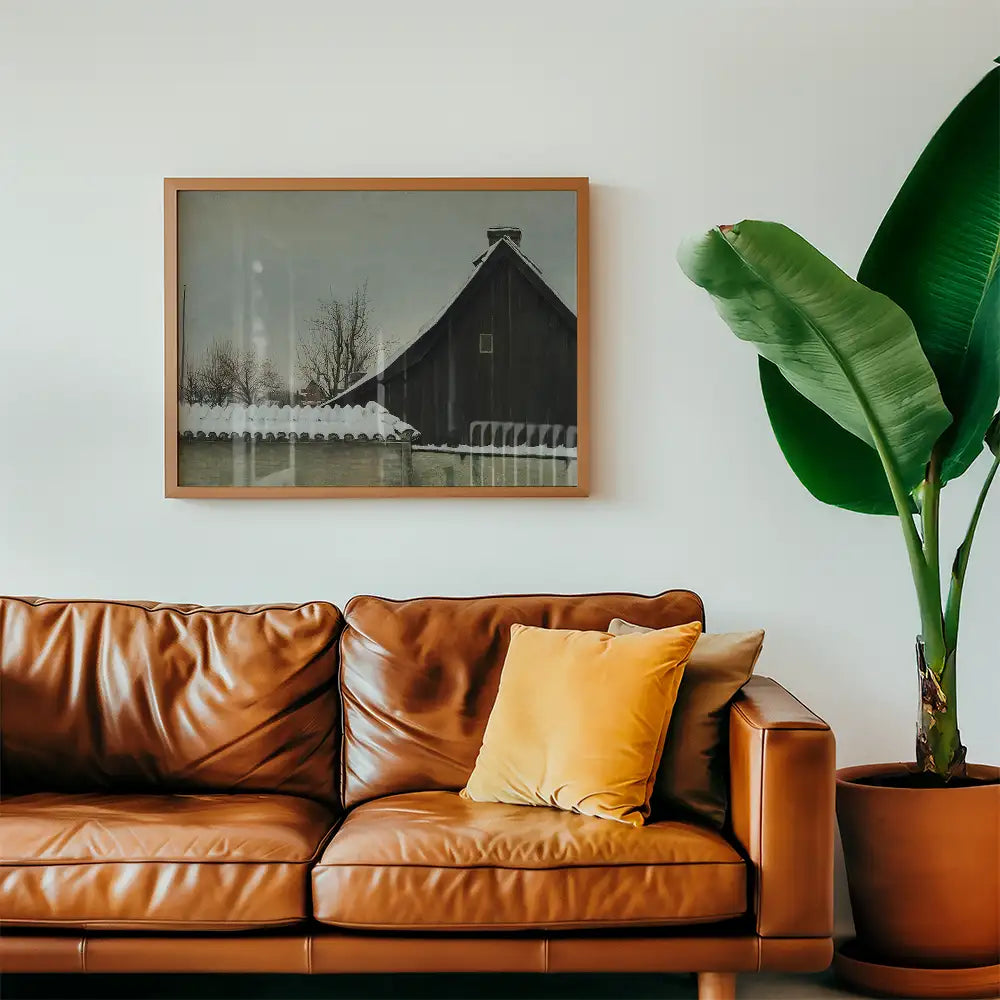 Framed painting of a winter landscape featuring a dark wooden barn and bare trees, displayed above a brown leather sofa with a yellow pillow, next to a large green plant.