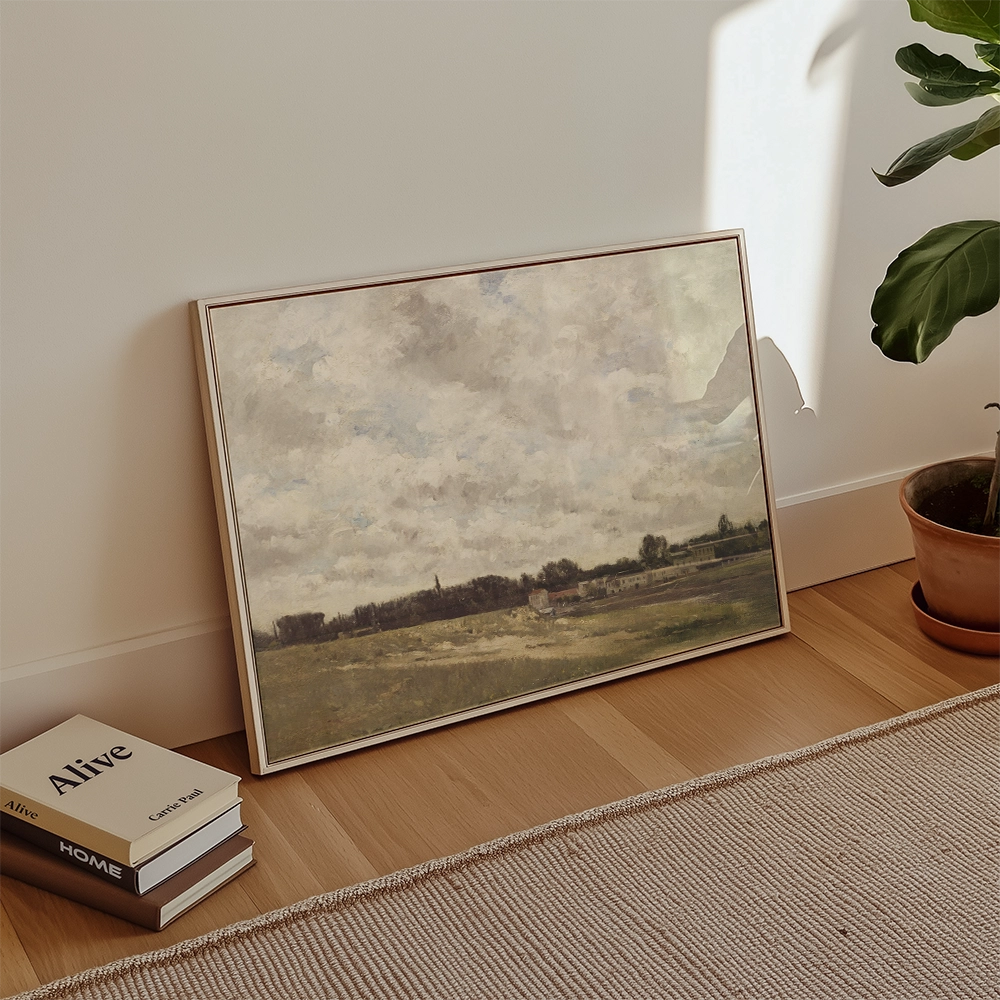 Framed landscape painting depicting a cloudy sky and fields, leaning against a wall beside a plant and books.