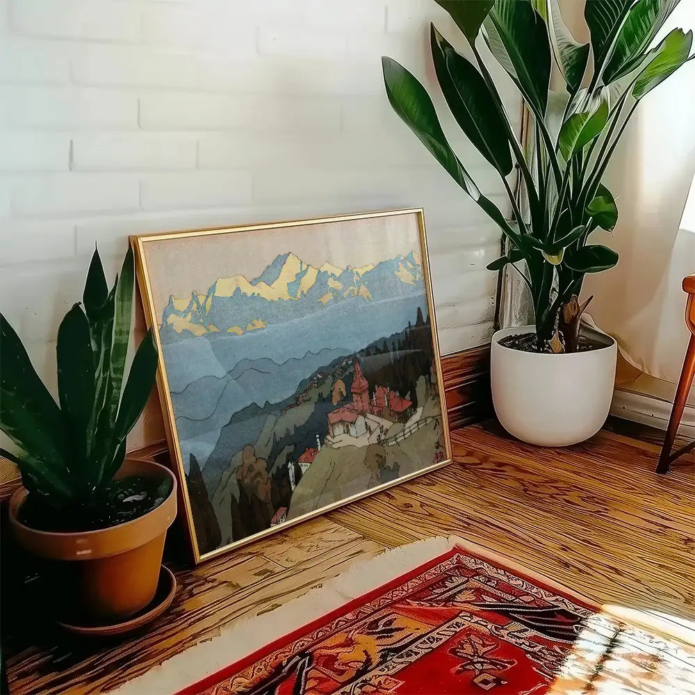 Framed landscape painting featuring mountains and a village, displayed next to a potted plant in a cozy indoor setting.