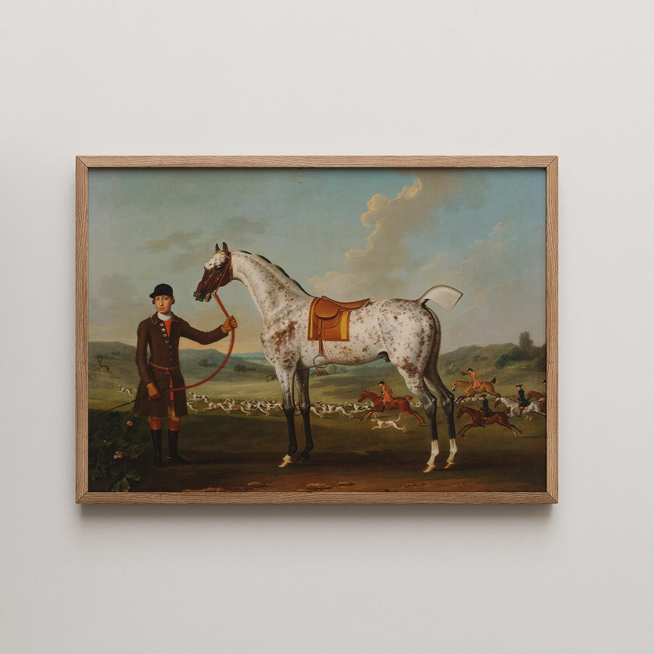 Painting of a man in a riding outfit holding a horse with a saddle, set in a landscape with a hunt scene in the background, framed in wood.