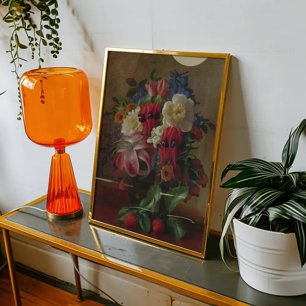 Framed floral painting displayed on a console table beside an orange lamp and a potted plant.