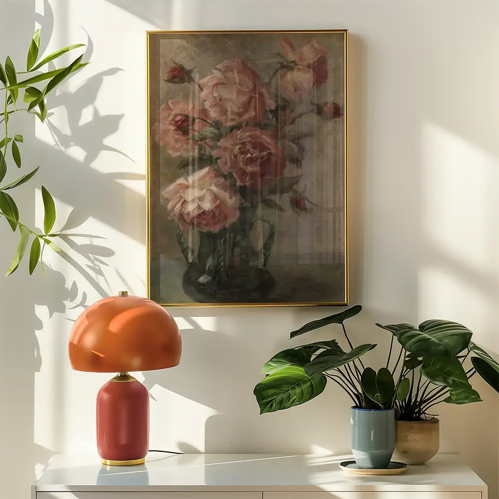 Painting of pink roses in a vase framed in gold, displayed on a wall beside a modern orange lamp and potted plants.