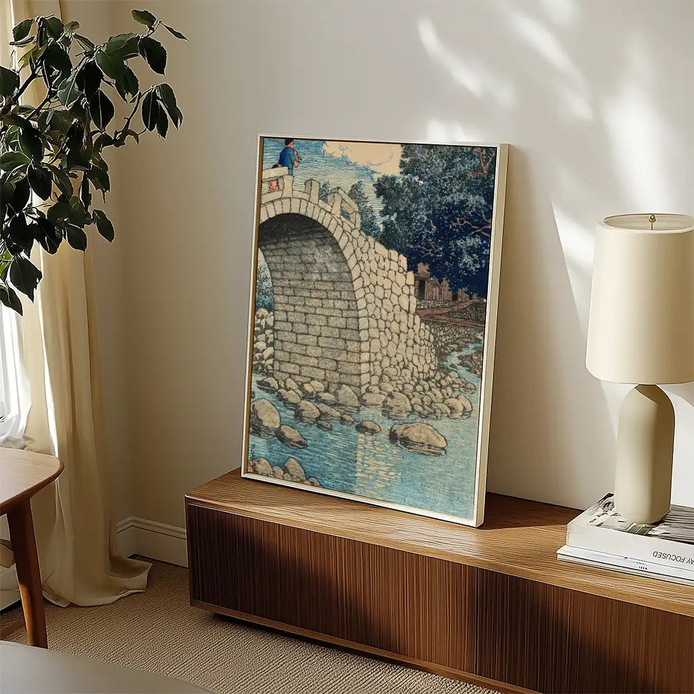 Framed painting of a stone bridge over a river with a person standing on the bridge, displayed in a living room setting with natural light and a lamp.