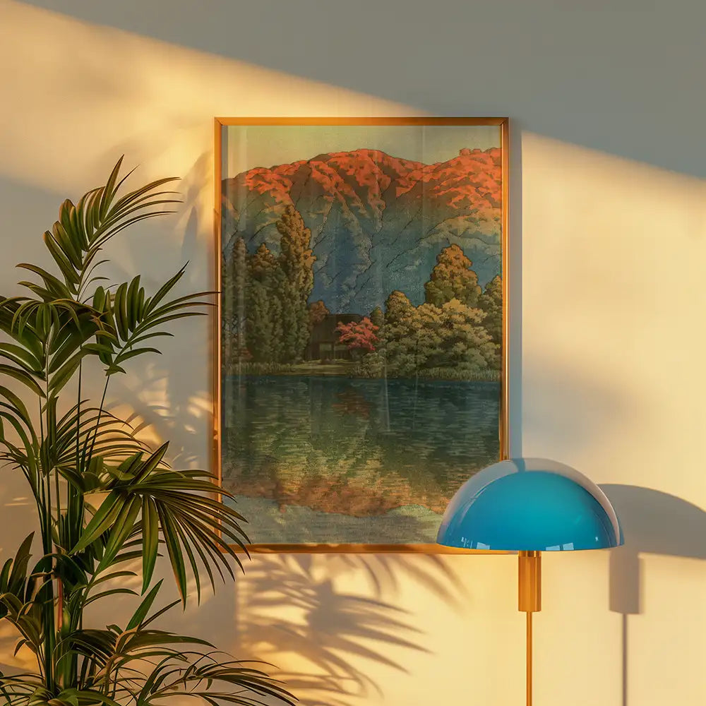 Framed painting of a serene landscape with mountains, trees, and a reflective lake, displayed on a wall next to a plant and a blue lamp.