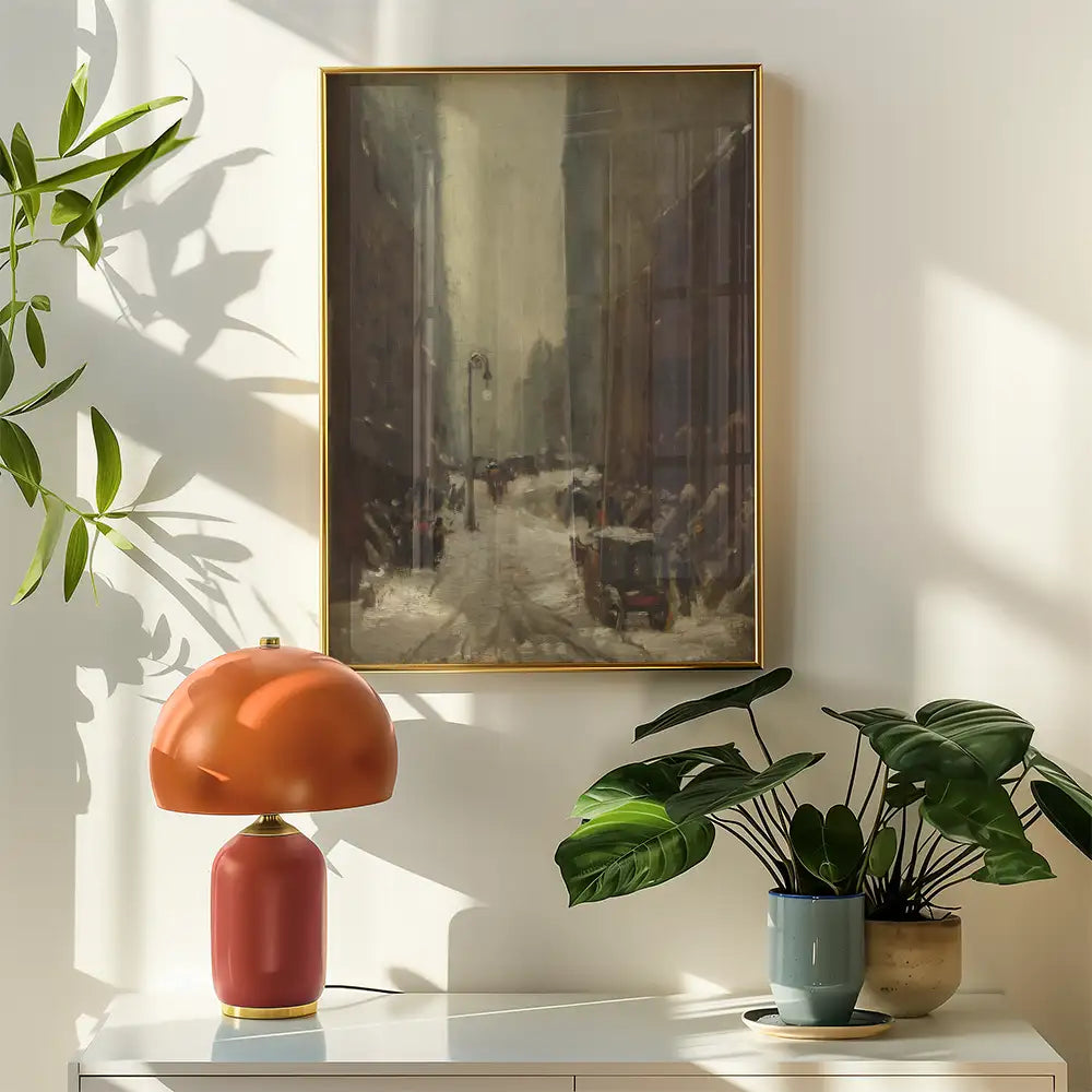 Painting of a snowy city street framed in gold, displayed above a modern orange lamp and potted plants on a white table.