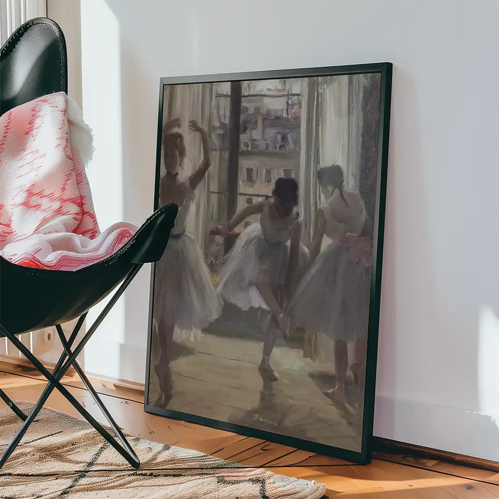 Framed painting of three ballerinas in a studio, showcasing soft colors and light filtering through a window, with a black chair and cozy blanket nearby.