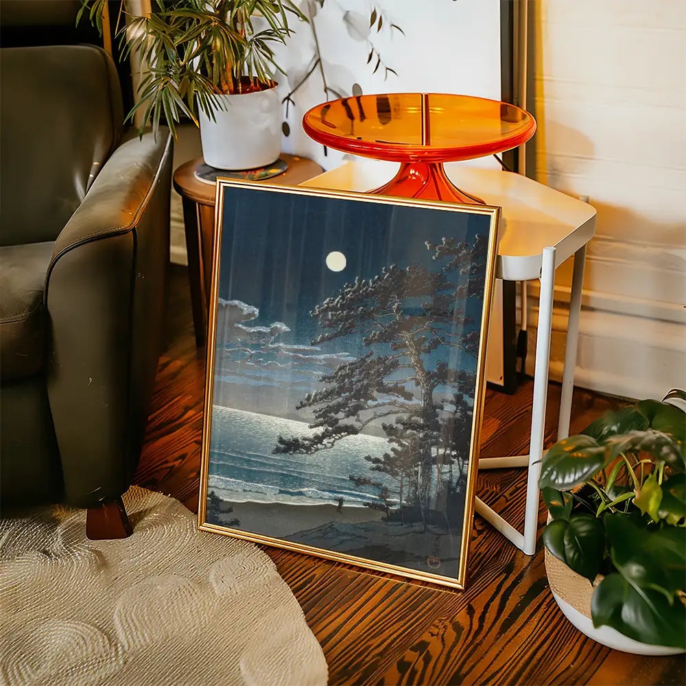 Framed painting of a moonlit seascape with trees in the foreground, placed next to a plant and a modern side table in a cozy living room setting.