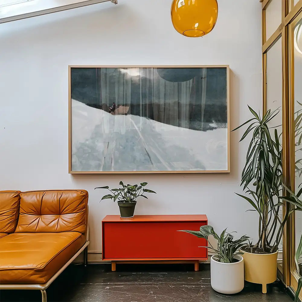 A large abstract painting in a wooden frame hanging on a white wall, above a red cabinet and surrounded by indoor plants, with a brown leather sofa in the foreground.