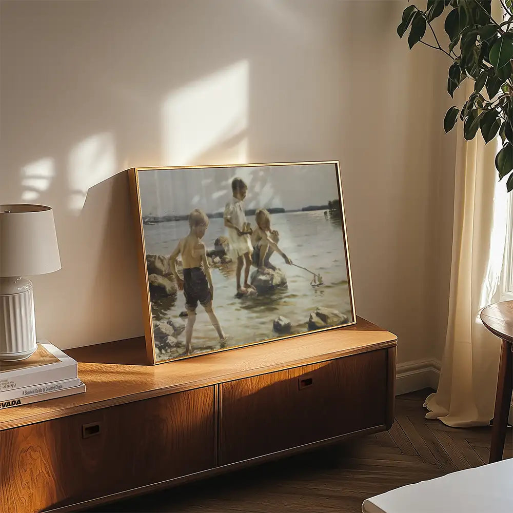 Painting of three children playing by the water, framed and displayed on a wooden surface in a cozy interior setting.