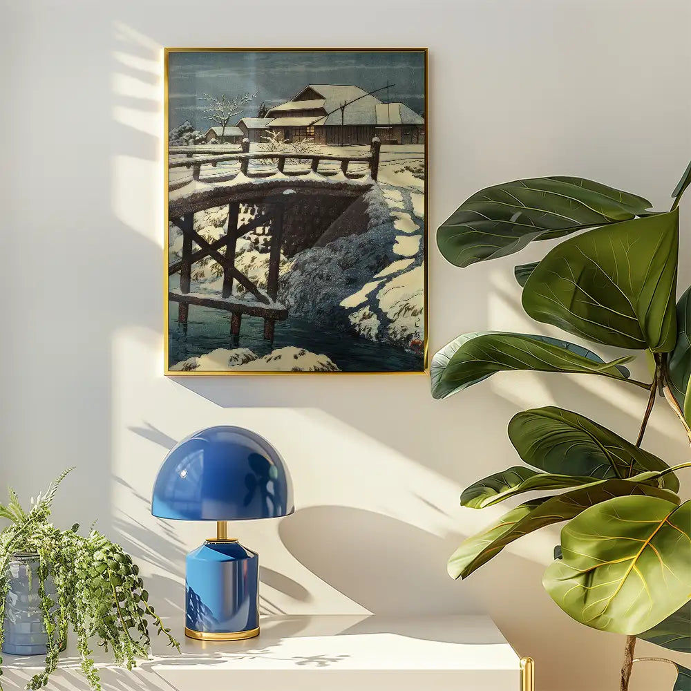 Painting of a snowy landscape featuring a bridge over a river, framed and hung on a wall near a blue lamp and indoor plants.