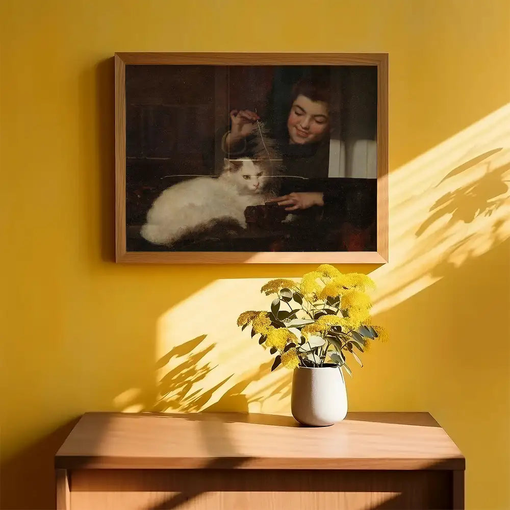 A framed painting of a girl playing with a white cat, hung on a bright yellow wall, with a vase of yellow flowers on a wooden table in the foreground.