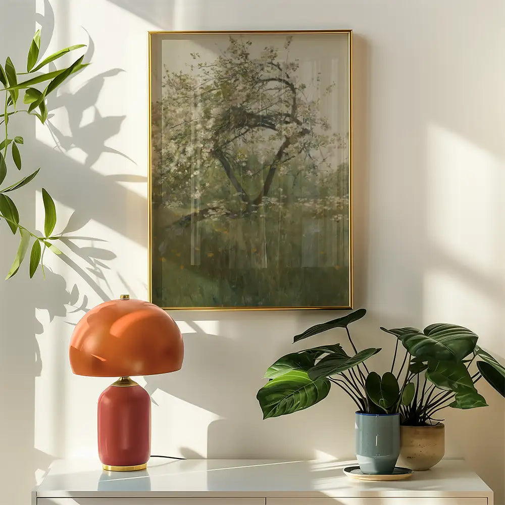 Painting of a tree in a soft, muted color palette, framed in gold, displayed above a stylish orange lamp and green potted plants on a white surface.