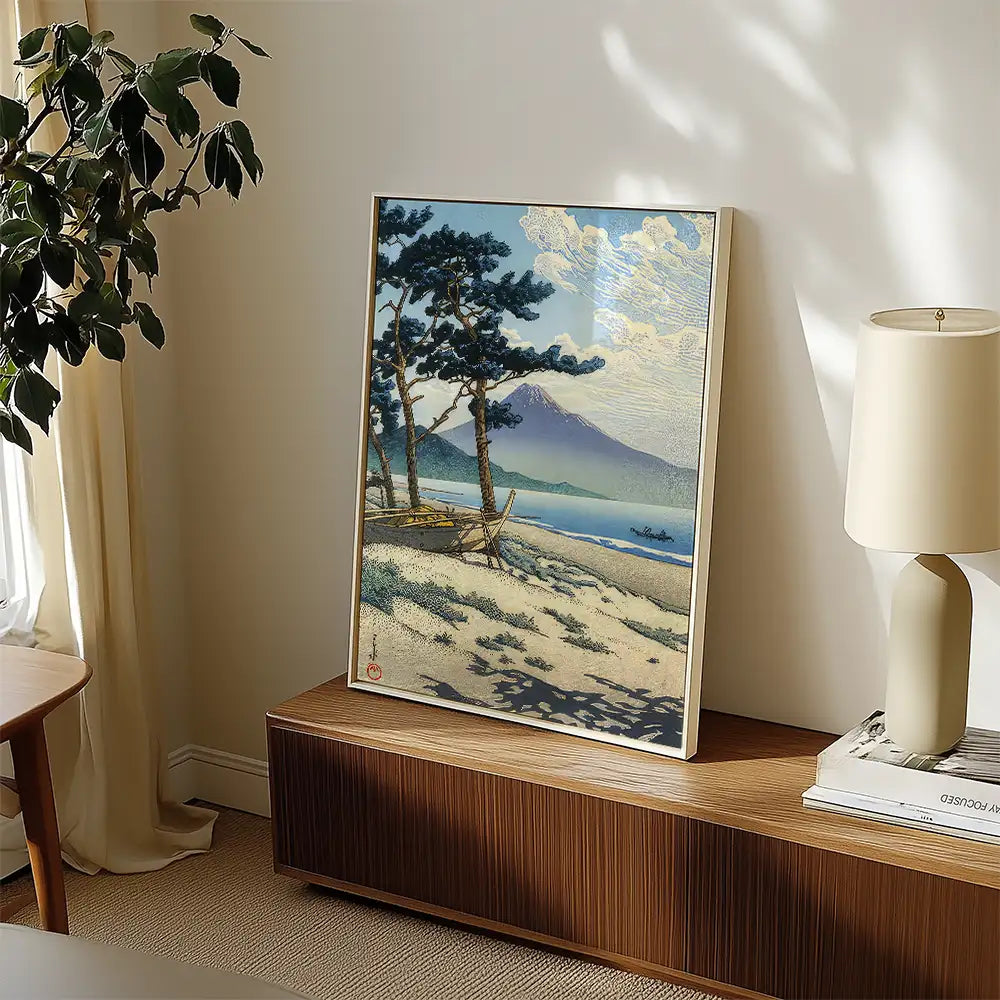 Framed landscape painting featuring a beach scene with trees and mountains, displayed in a modern interior setting.