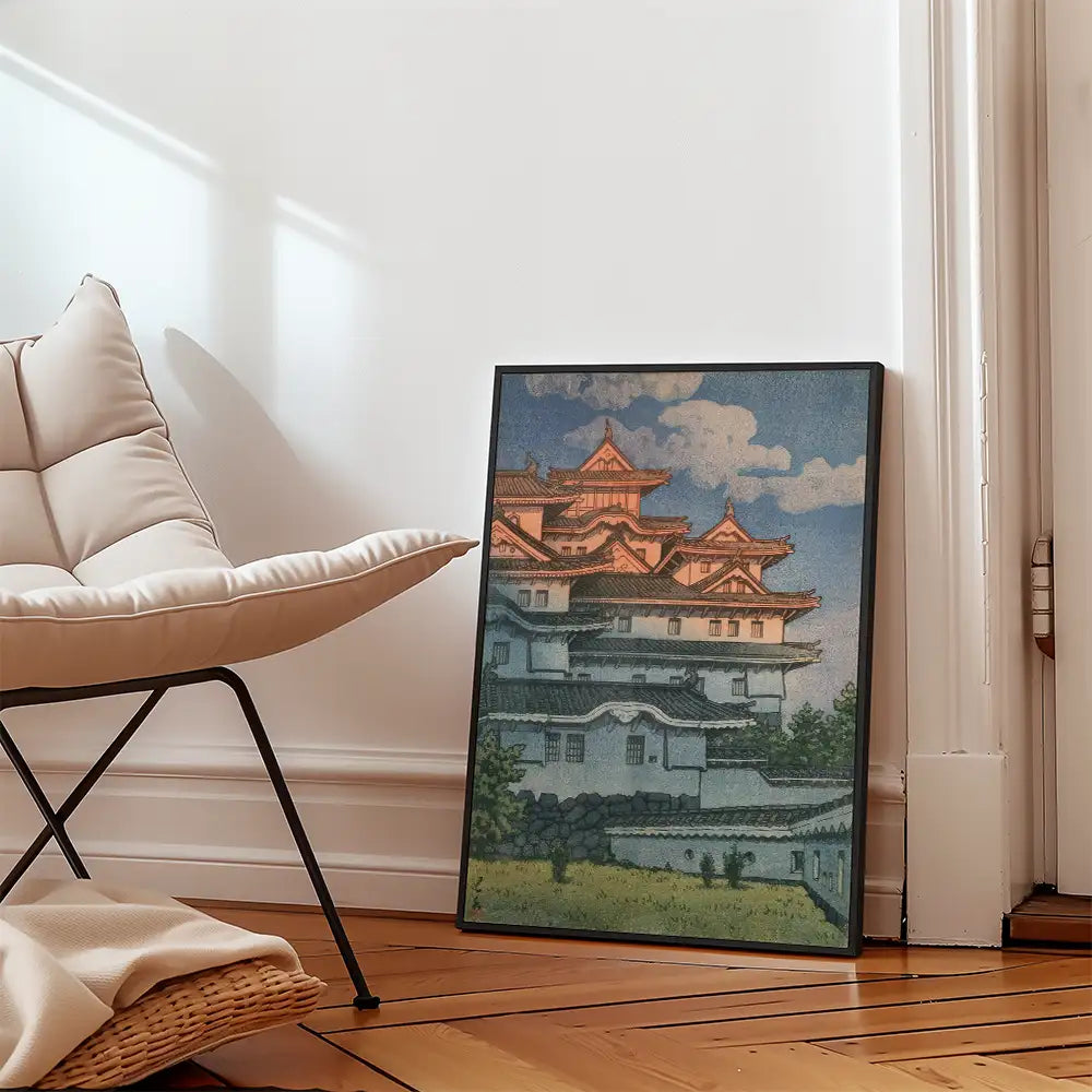 Framed painting of a traditional Japanese castle against a cloudy sky, leaning against a wall in a modern interior setting with a contemporary chair.