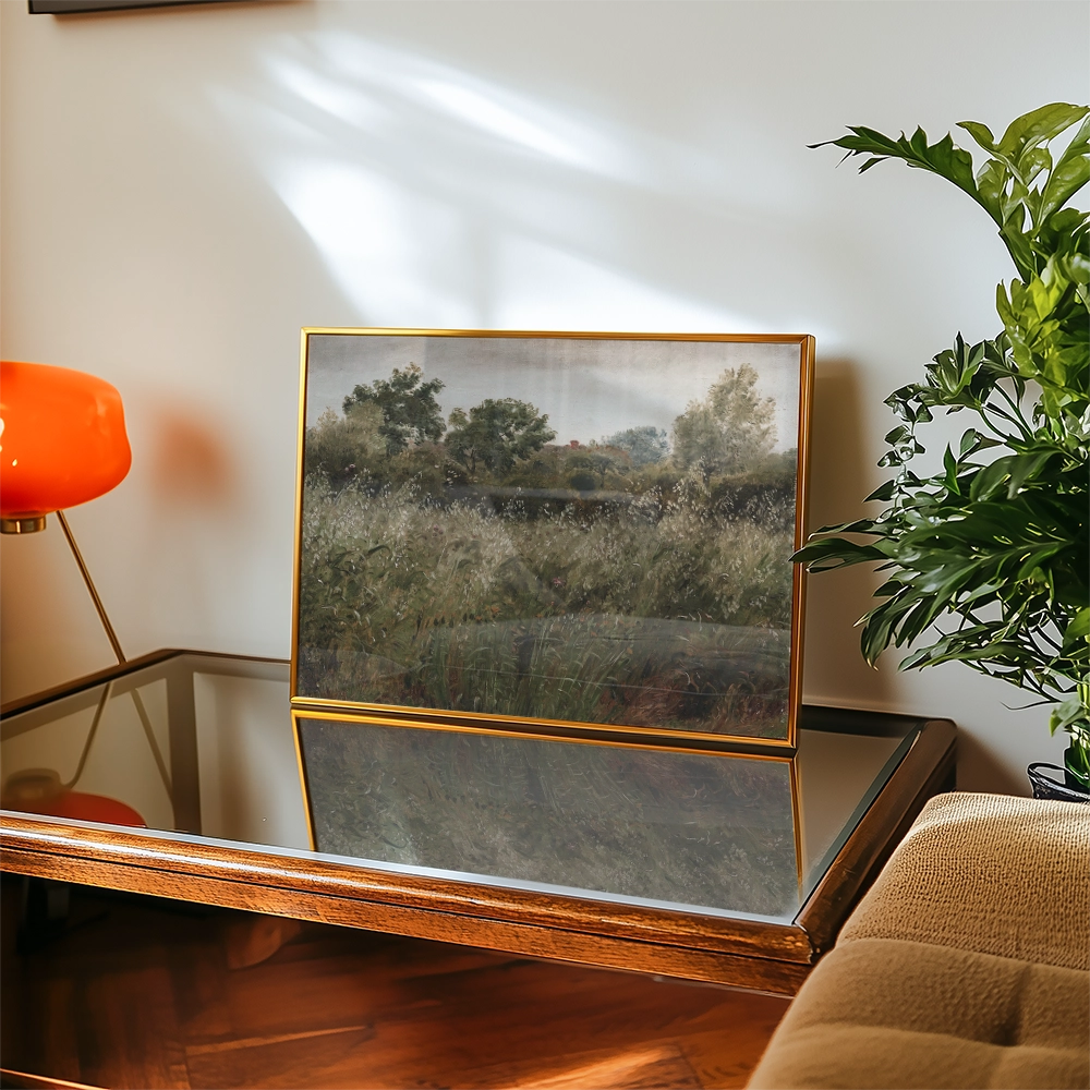 Painting of a landscape featuring trees and grasses, displayed in a gold frame on a wooden table with a glass surface, alongside a potted plant and an orange lamp.