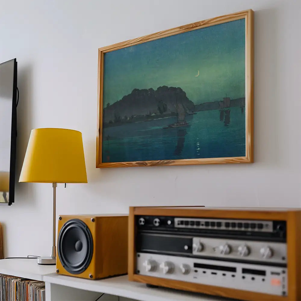 Framed painting of a serene landscape with a crescent moon over a body of water, displayed on a wall near a yellow lamp and vintage speakers.