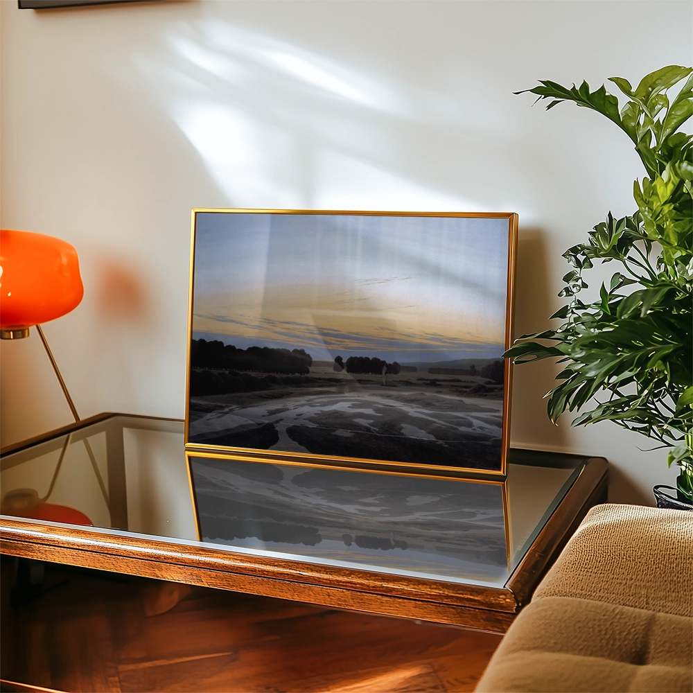 A framed landscape painting featuring a serene view of trees and a sunset, displayed on a reflective coffee table beside an orange lamp and greenery.