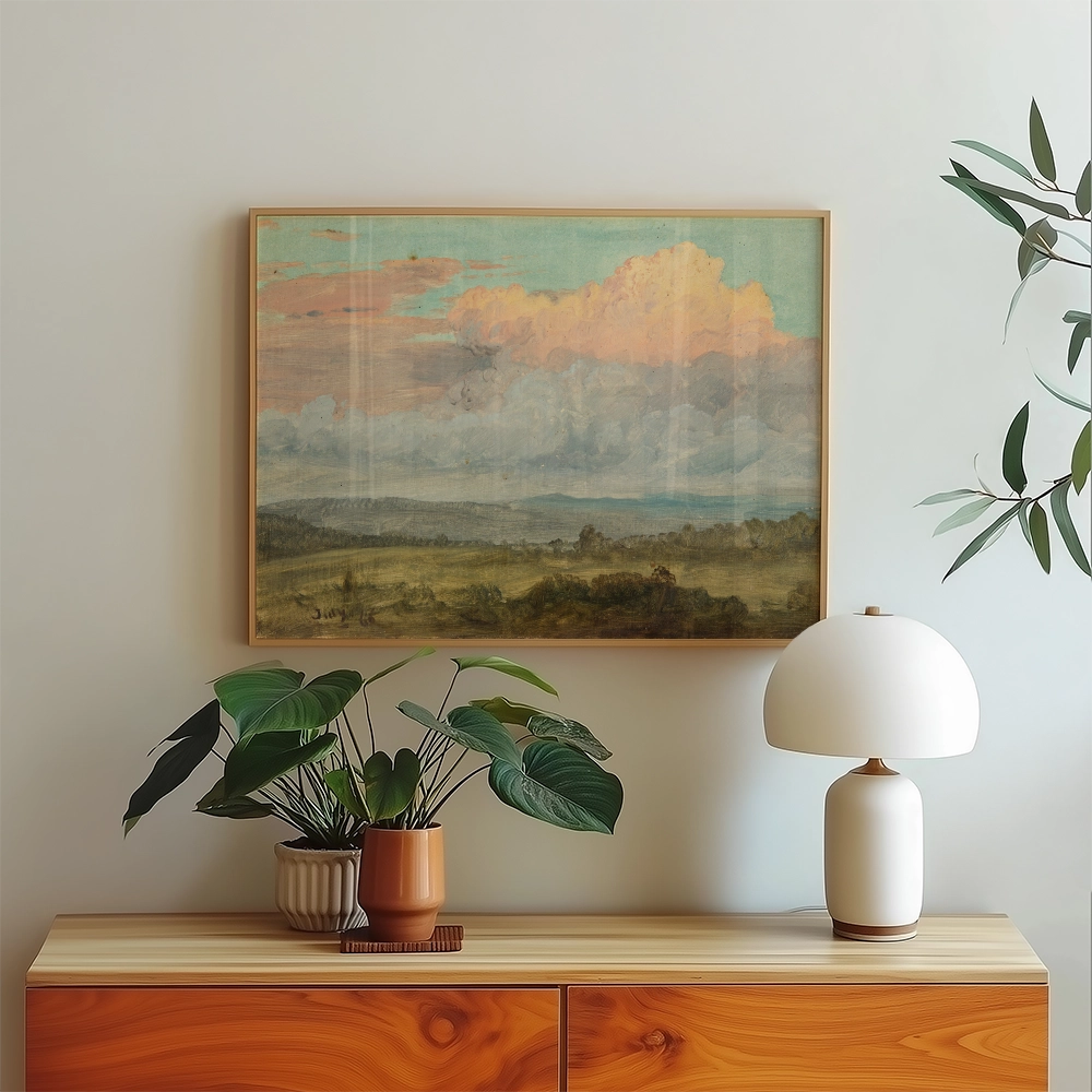 Framed landscape painting with clouds above a green field, displayed on a light wooden cabinet alongside a potted plant and a modern lamp.