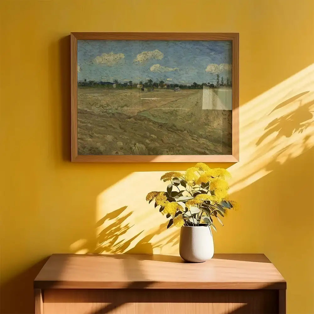Framed landscape painting on yellow wall with sunlight casting shadows, accompanied by a vase of yellow flowers on a wooden side table.